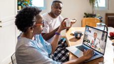 Person on laptop at kitchen table with another who holds up prescription bottle for telehealth doctor 