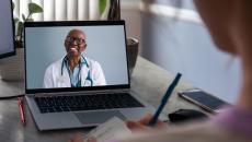 Female doctor on laptop smiles at patient in foreground who is taking notes