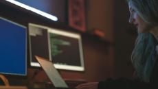 Person with long hair sits before a laptop and multiple screens