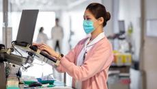 Female nurse in mask and pink sweater types notes in a hospital hallway