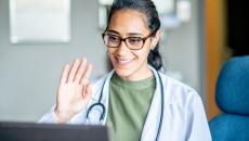 Female doctor smiles and waves into a laptop during a telehealth visit