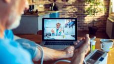 Male doctor appears on screen as patient in foreground checks his blood pressure at home.