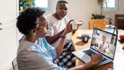 Person on laptop at kitchen table with another who holds up prescription bottle for telehealth doctor 