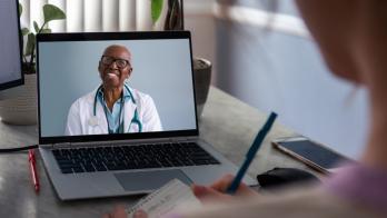 Female doctor on laptop smiles at patient in foreground who is taking notes