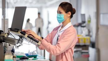 Nurse in mask and pink sweater types notes in a hospital hallway