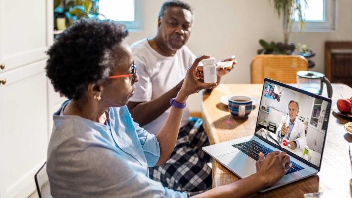 Person on laptop at kitchen table with another who holds up prescription bottle for telehealth doctor 